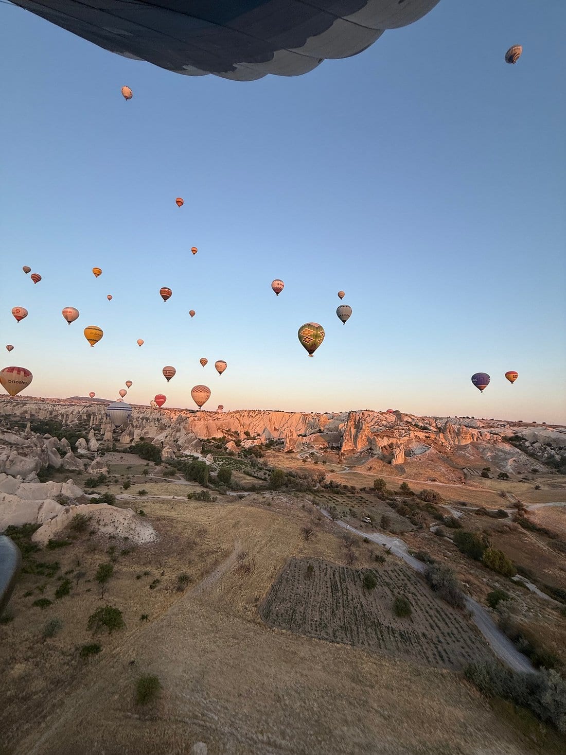 La vista dall’interno della cesta di una mongolfiera guardando in basso verso decine di mongolfiere sulla valle della Cappadocia all’alba