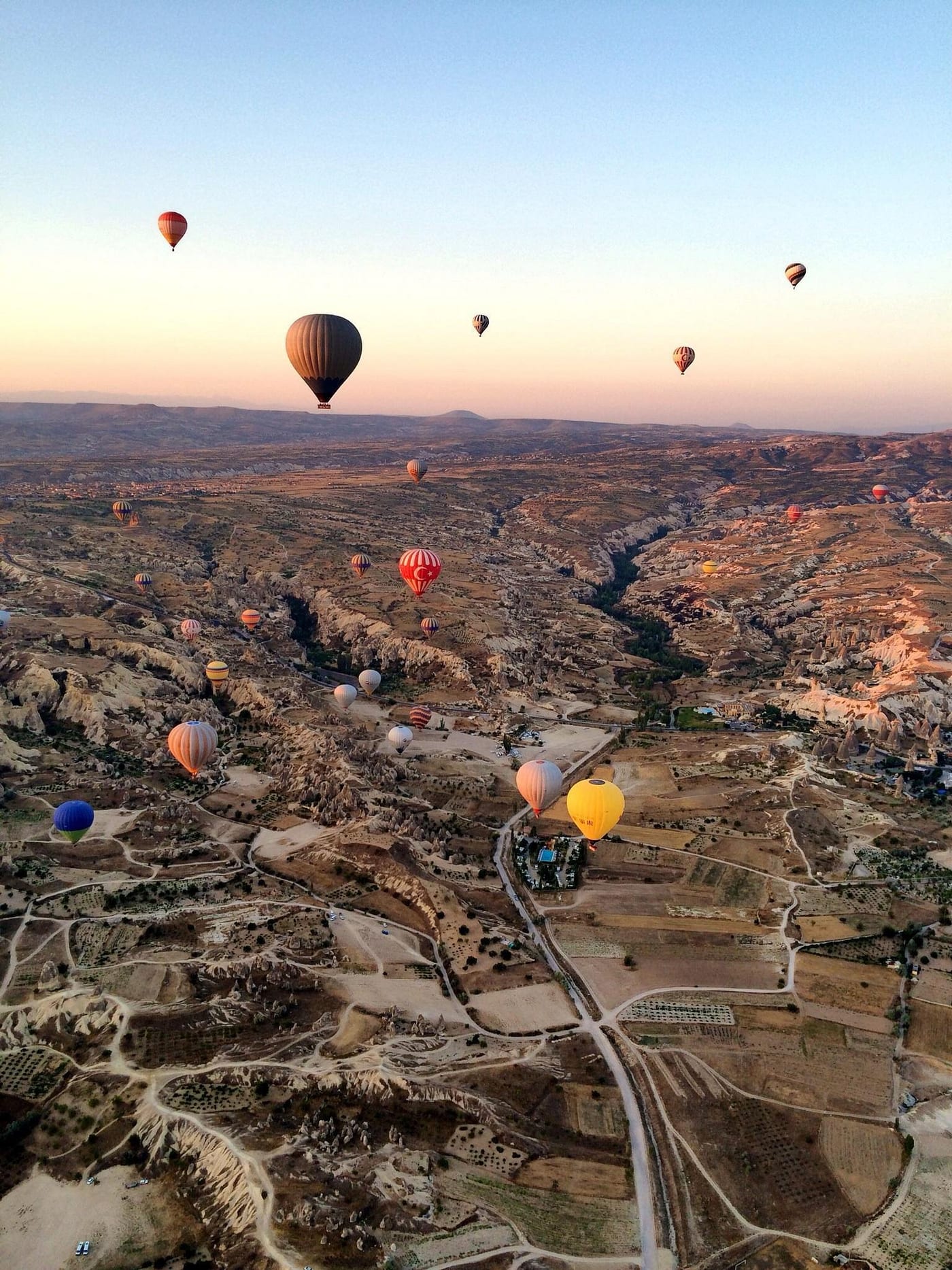 Vista aerea dall’alto sui campi a mosaico e sulle valli della Cappadocia con mongolfiere che scivolano ad altezze diverse