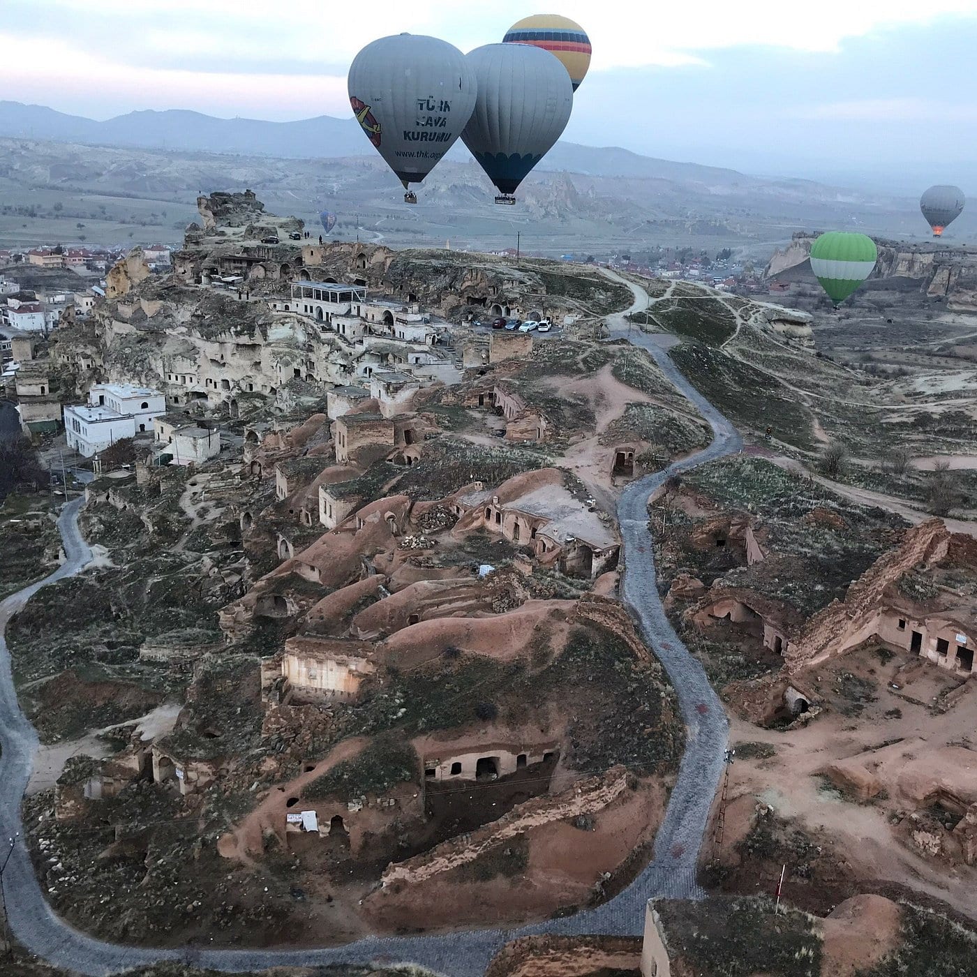 Vista aerea di un paese scavato nella roccia della Cappadocia con mongolfiere che scivolano sopra la cresta alle spalle