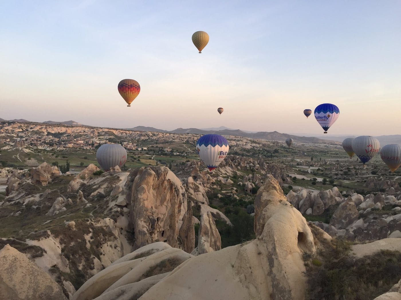 Vista ampia di mongolfiere che si alzano sui camini delle fate e sulle abitazioni rupestri di Göreme all’alba
