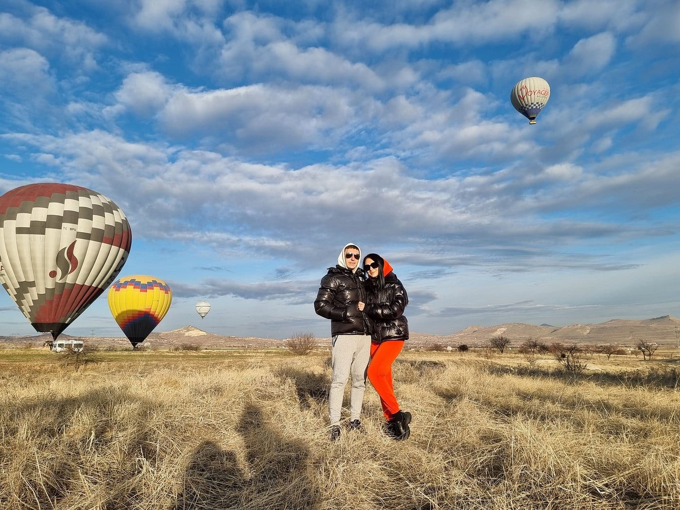 Una coppia con cappotti invernali in piedi su un campo di decollo in Cappadocia con mongolfiere che si alzano nel cielo azzurro del mattino