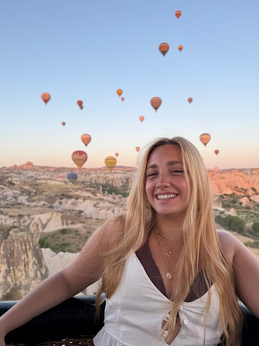 Un’ospite sorridente nella cesta di una mongolfiera con decine di mongolfiere che si alzano sulla valle della Cappadocia all’alba