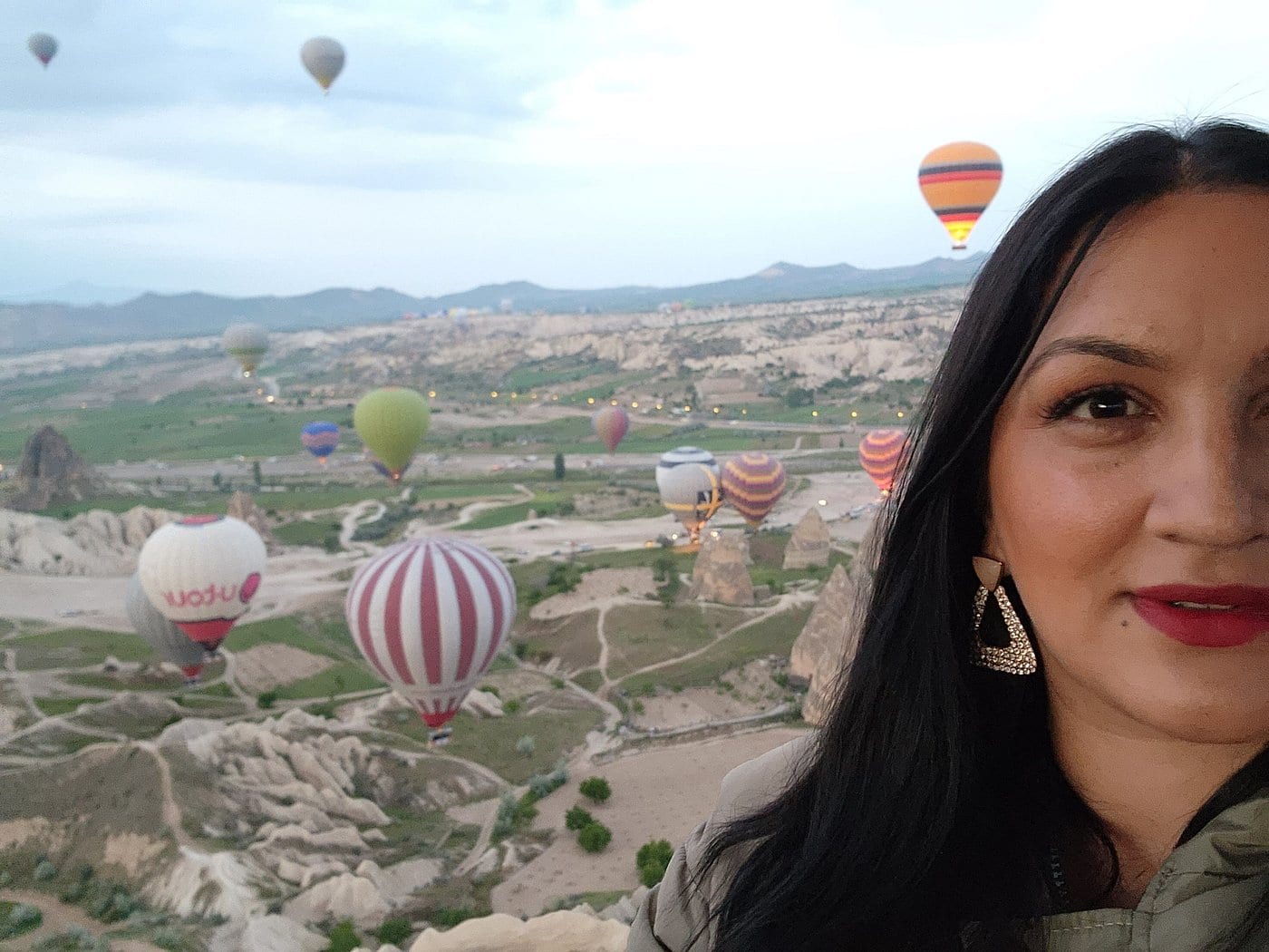 Un’ospite che si volta verso la fotocamera nella cesta di una mongolfiera con la valle della Cappadocia e le mongolfiere alle spalle all’alba