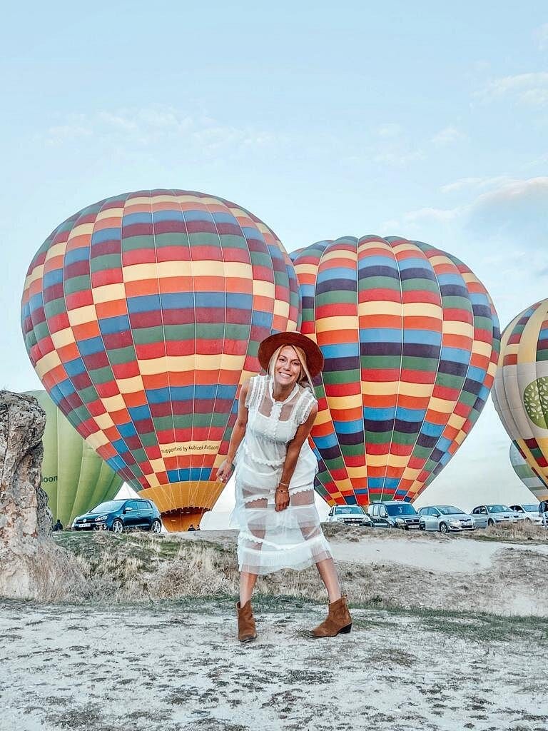 Un’ospite con abito bianco e cappello davanti a mongolfiere colorate che si gonfiano su un campo di decollo in Cappadocia