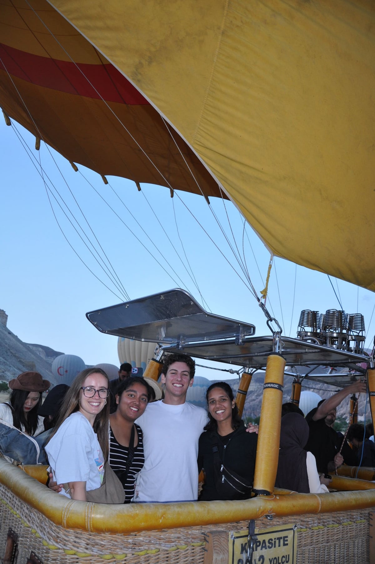 Four guests smiling together in a Comfort hot air balloon basket over Cappadocia, with the burners and other balloons behind them