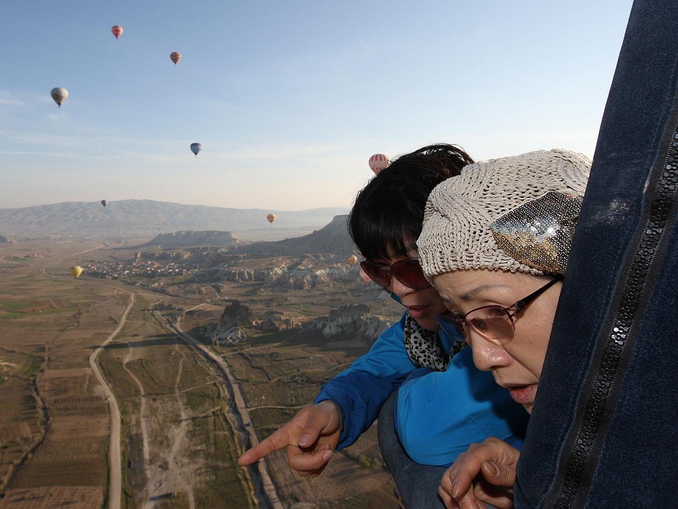 Due ospiti affacciate al bordo della cesta di una mongolfiera, una che indica la valle della Cappadocia in basso