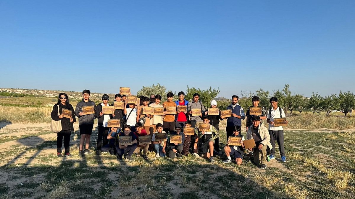 A large group of Hot Air Cappadocia Balloon guests gathered after their flight, each holding a personalized flight certificate, captured in the open landing field with the Cappadocia landscape behind