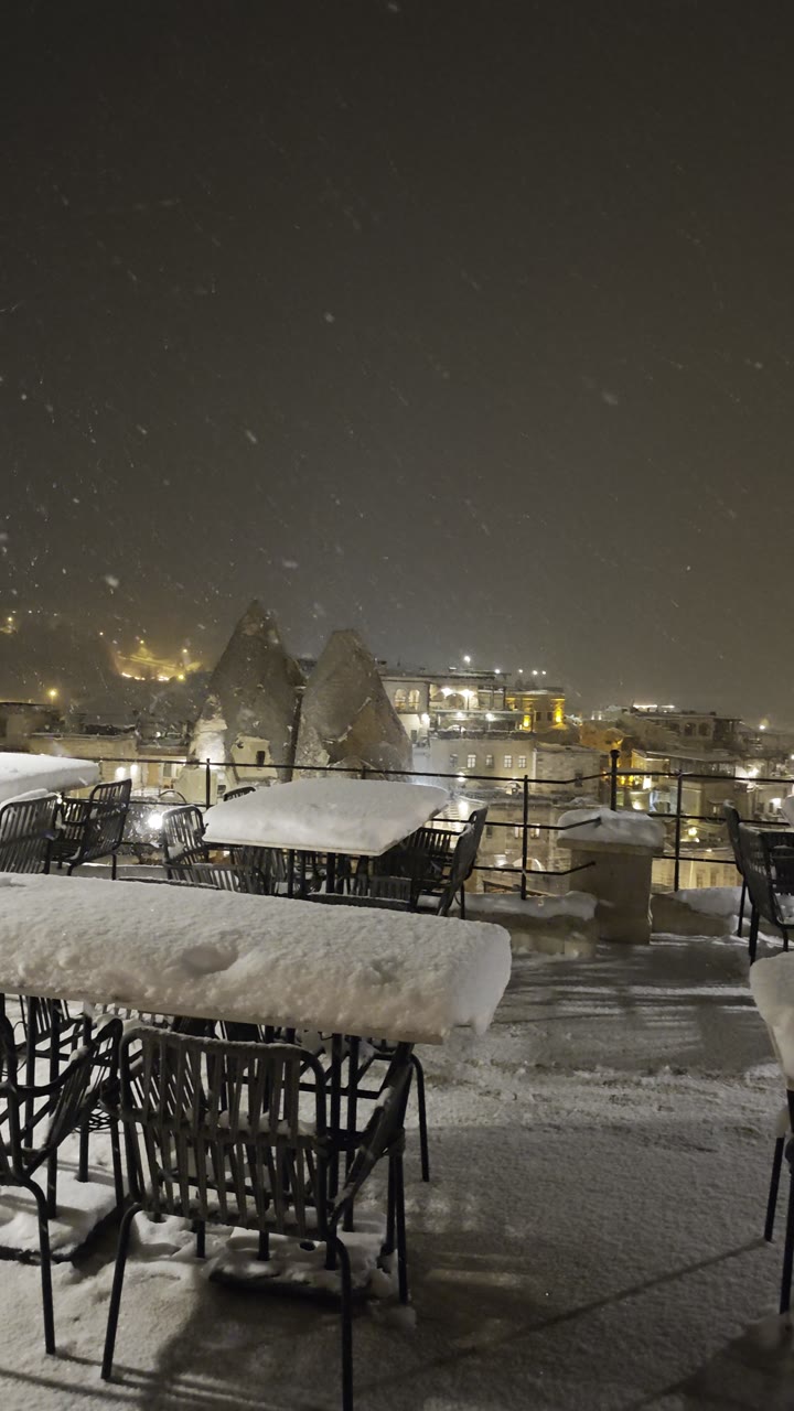 Neve che cade di notte sulla terrazza di un hotel nella roccia a Göreme, con i camini delle fate e le luci del paese dietro