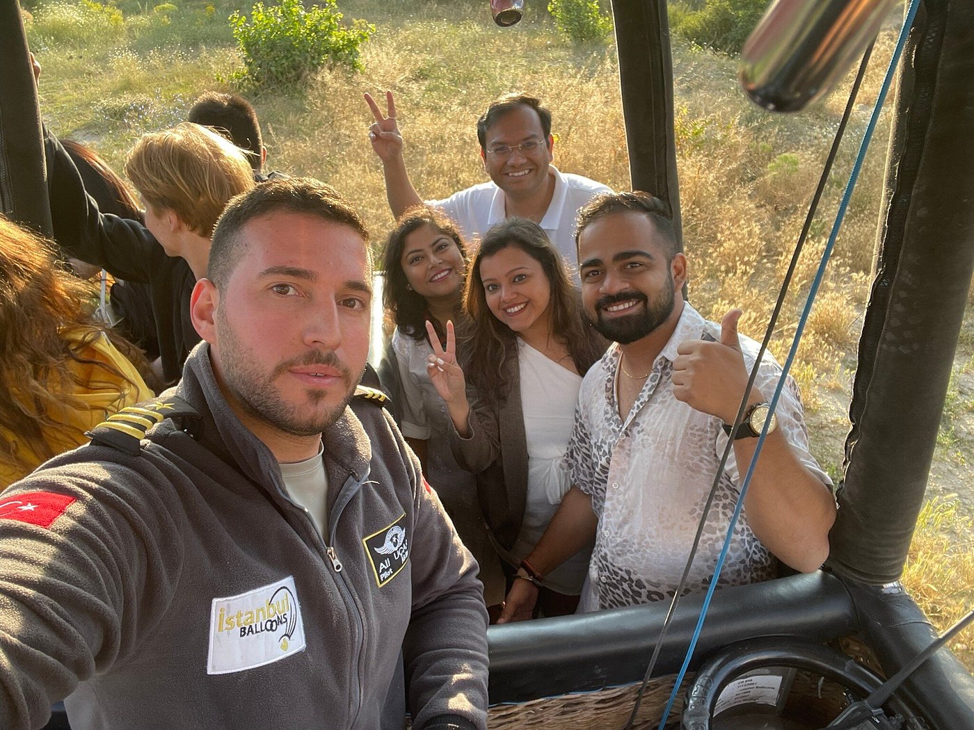 A pilot and first-time guests in a hot air balloon basket at sunrise over Göreme, Cappadocia, with dozens of balloons rising across the valley