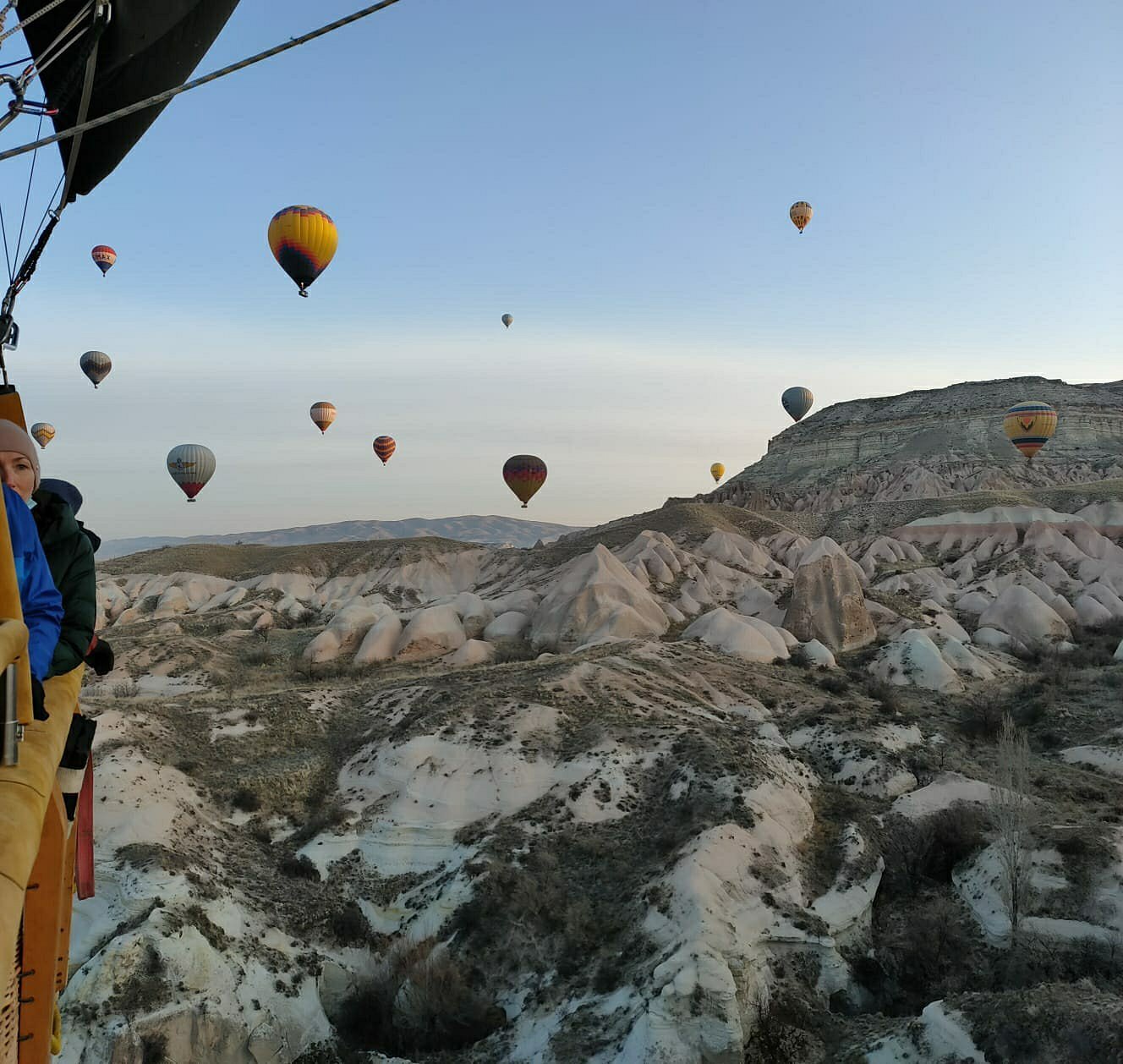 A 28-passenger hot air balloon flies low over the Cappadocia countryside, with smaller balloons in the background