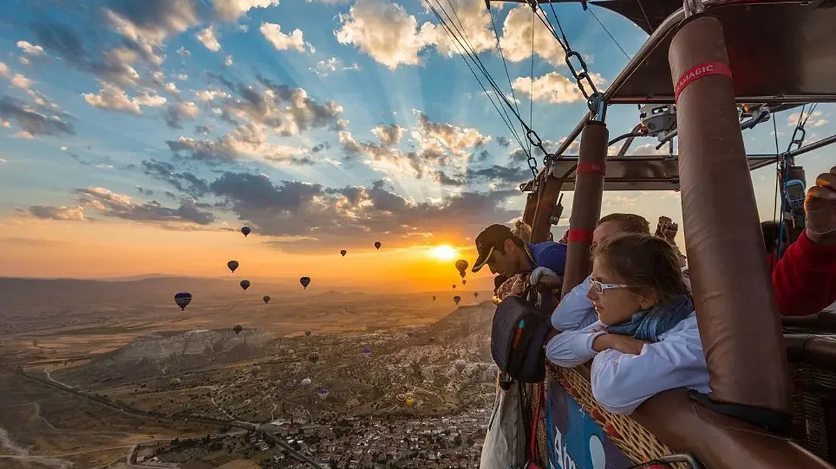 Passengers in a hot air balloon basket watching the sunrise over Cappadocia, with other balloons drifting above the valley
