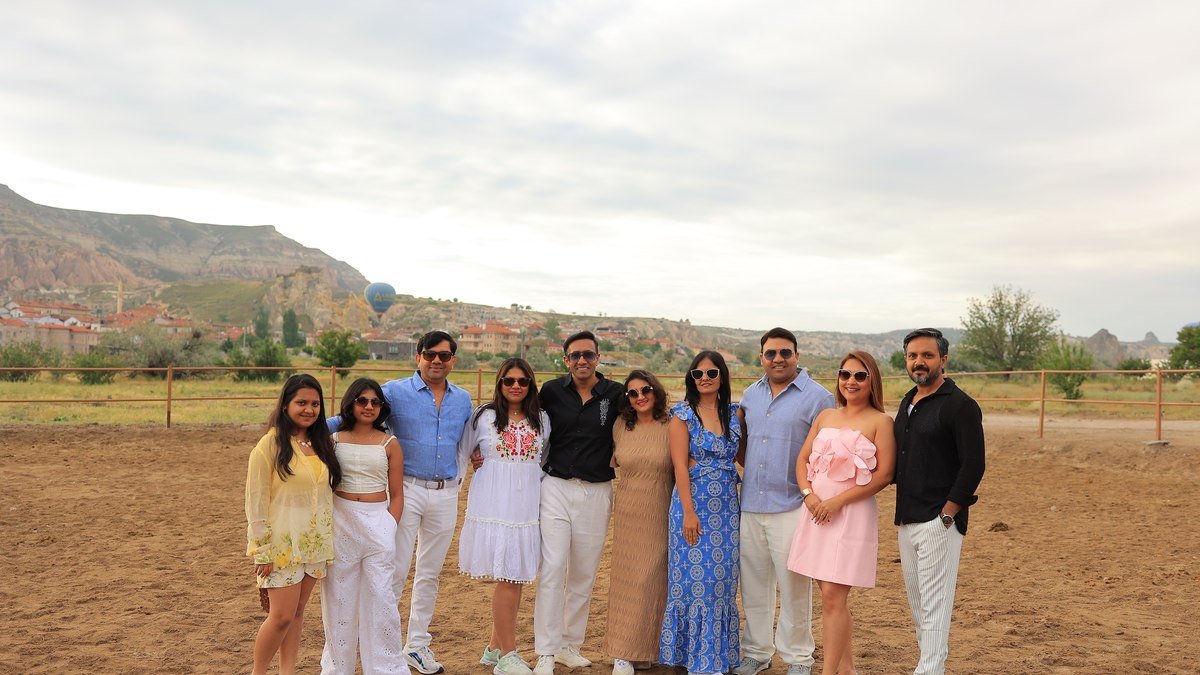 A family group of guests standing together in the Cappadocia landscape, with a hot air balloon in flight and the volcanic hills and village behind them, capturing the combination of a balloon morning and a Cappadocia tour day