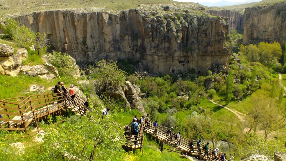 Visitors descending the wooden boardwalk staircase into Ihlara Valley, with the deep canyon walls of the Melendiz River gorge rising above them, the central walking section of the Cappadocia Green Tour