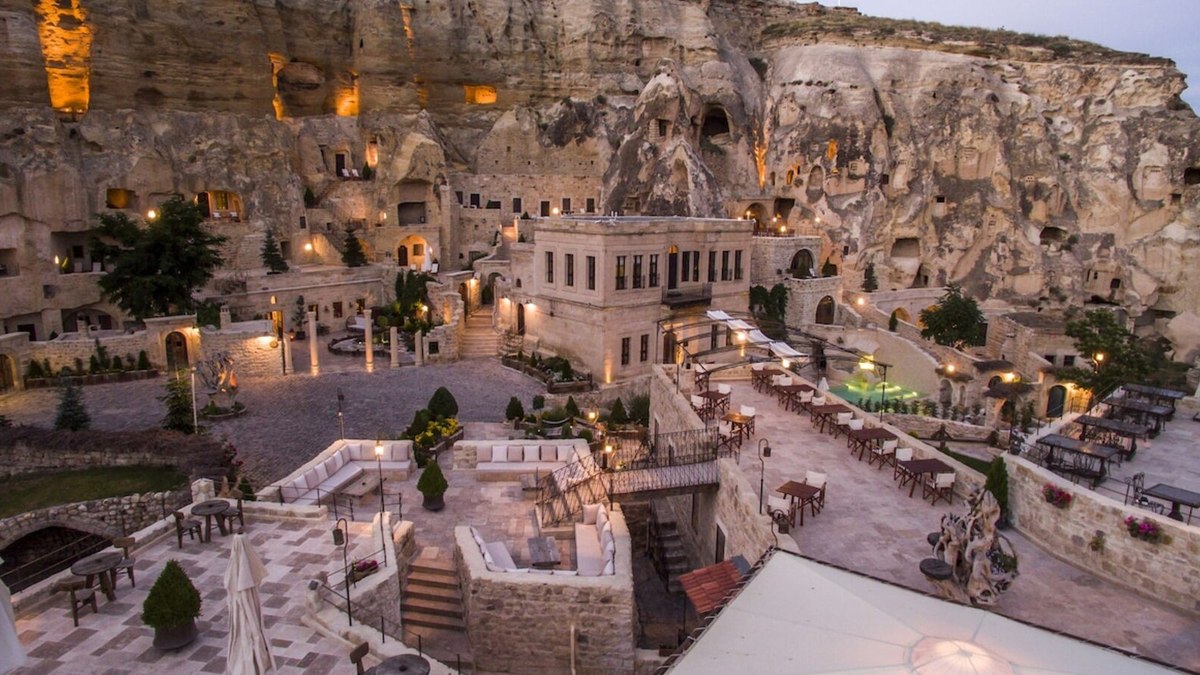 A restored stone courtyard of a boutique cave hotel in Ürgüp at twilight, with terraces, lit pathways and rooms carved into the rock face behind